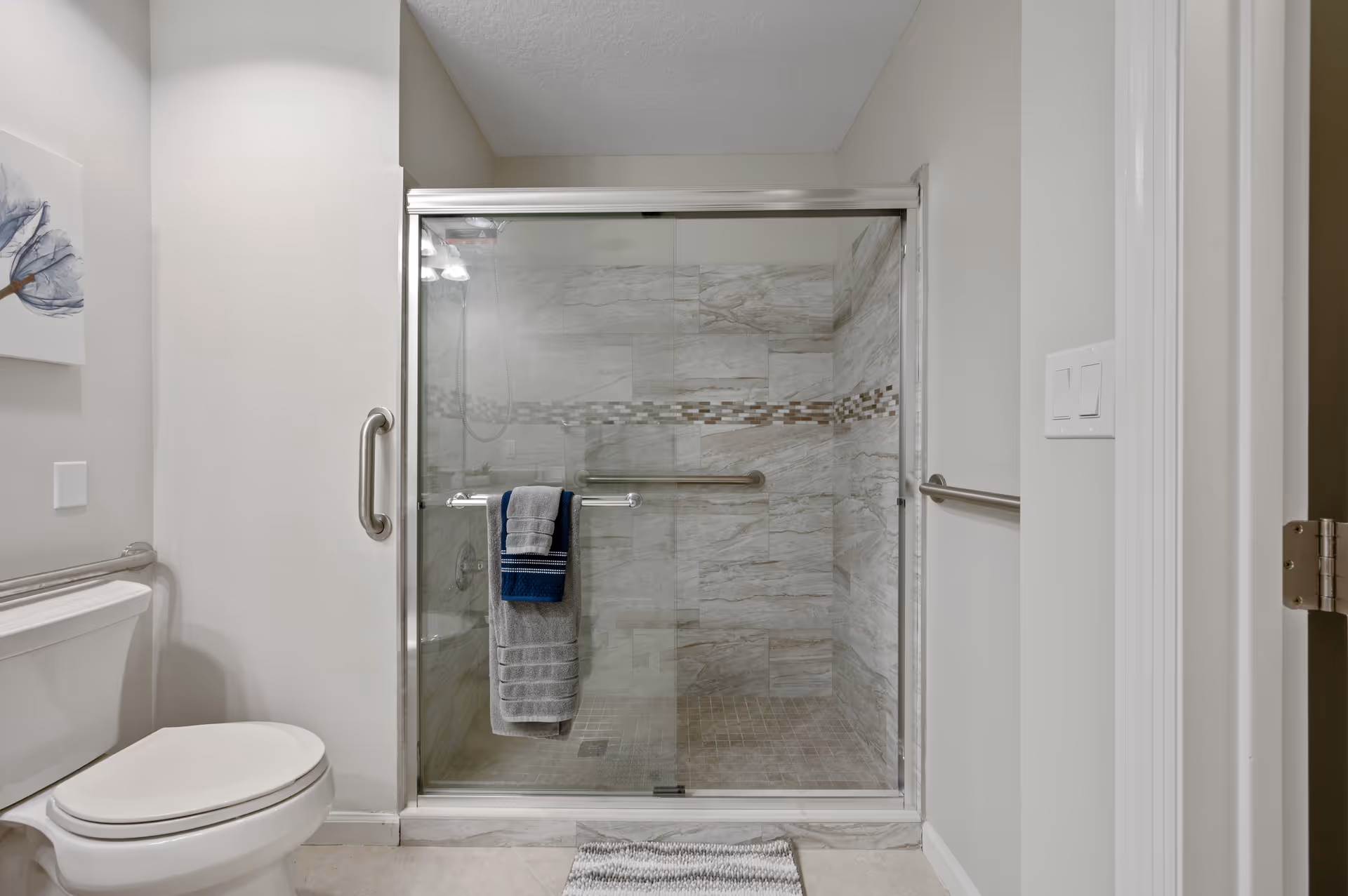 A modern bathroom featuring a glass-enclosed shower with beige and brown tiled walls and floor, a silver grab bar inside the shower, and two towels hanging on the shower door. To the left is a white toilet with a silver grab bar on the wall behind it. The walls are painted light beige and there is a small piece of artwork on the left wall.