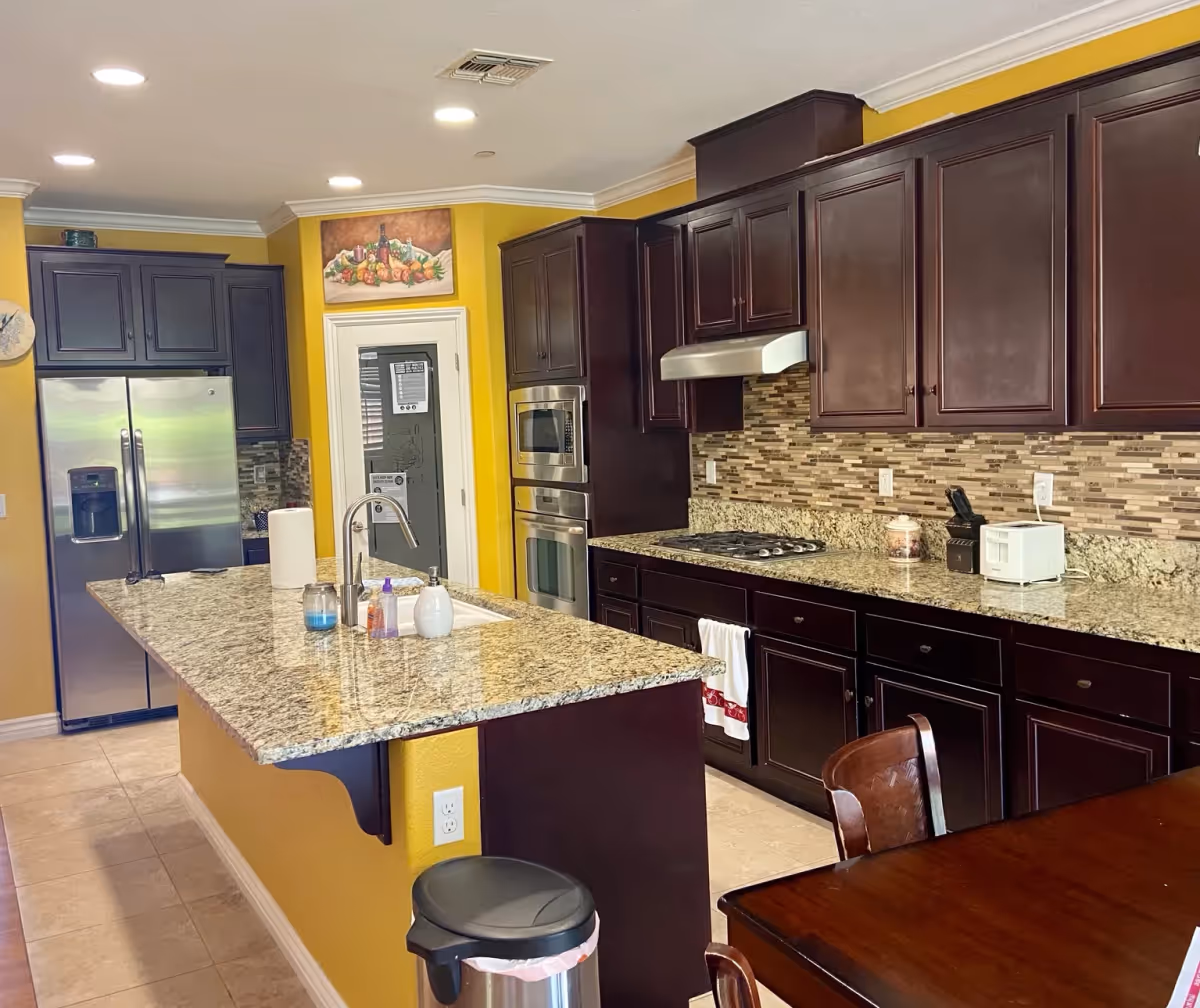 A modern kitchen with dark wood cabinets, granite countertops, and a tiled backsplash. The kitchen features a large island with a sink and faucet, a stainless steel refrigerator, built-in microwave and oven, a stovetop with a range hood, and various kitchen items including a toaster, knife block, and soap dispensers. The walls are painted yellow, and there is a dining table with chairs partially visible in the foreground.