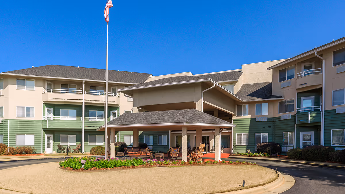 Exterior view of Asher Point Independent Living of Montgomery, showing a multi-story building with green and beige siding, a covered entrance with outdoor seating, a flagpole with an American flag, and a clear blue sky.