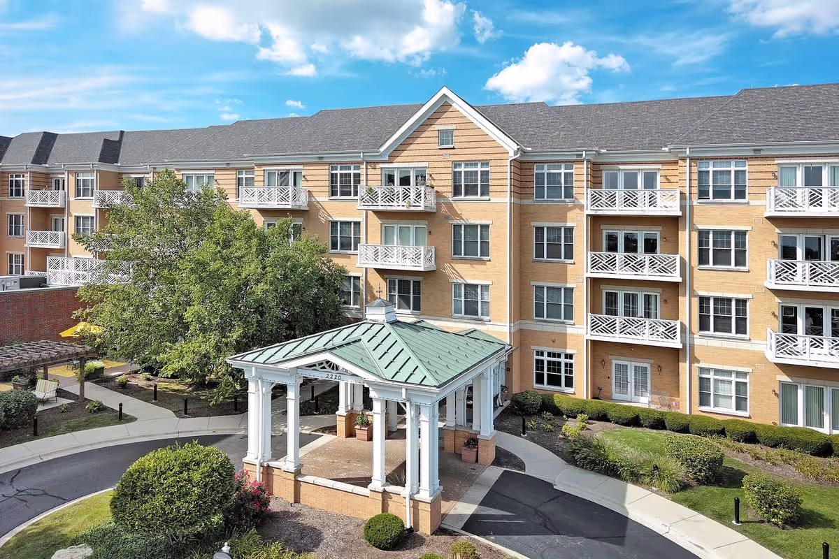 Exterior view of a multi-story senior living facility building with beige brick walls and white balconies. In front of the building is a covered entrance with white columns and a green roof, surrounded by landscaped greenery and a circular driveway. The sky is blue with some clouds.