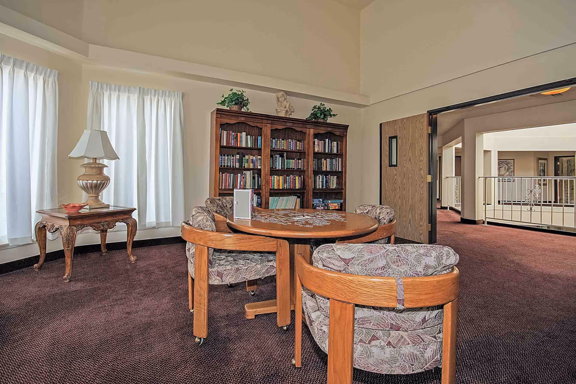 Small common room with a round wooden table and upholstered chairs, a bookshelf, side table with lamp, and curtained windows.