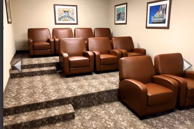 A small theater room with three rows of brown leather recliner chairs arranged on carpeted tiered steps. The walls are beige and decorated with framed pictures related to cinema and theater.