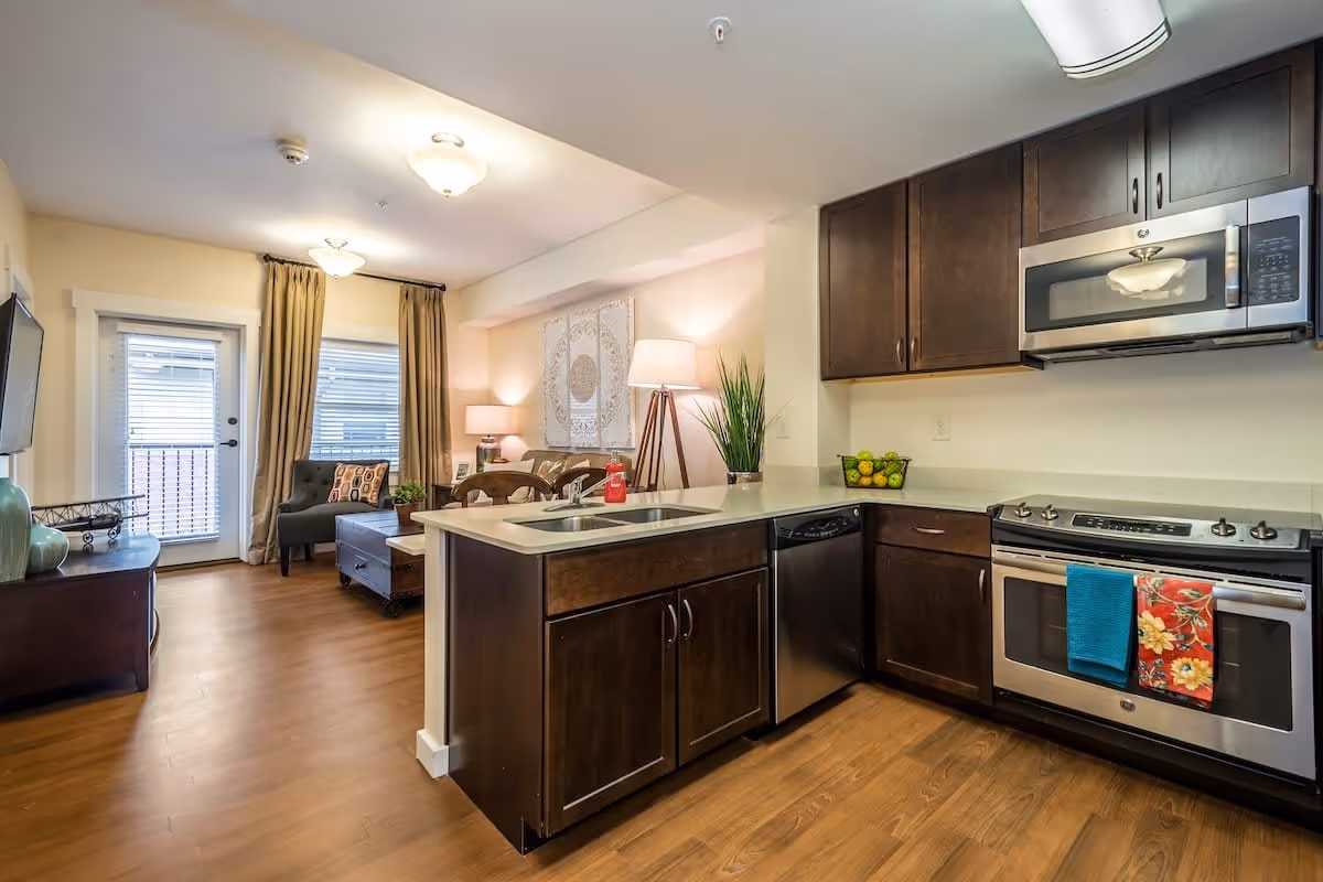 Interior view of a modern kitchen and living room area in a senior living facility. The kitchen features dark wood cabinets, a stainless steel microwave, oven, dishwasher, and a sink on a light-colored countertop. The living room has a chair, coffee table, TV on a stand, and large windows with beige curtains letting in natural light.
