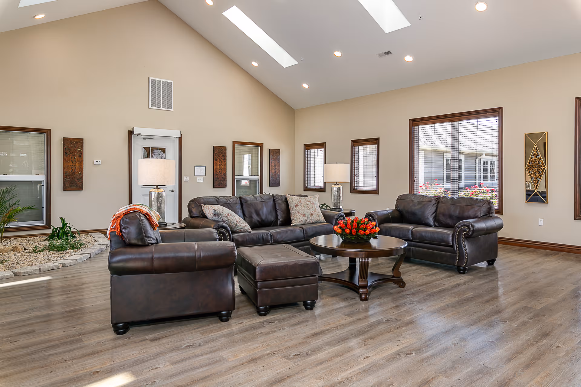 A spacious living room with high vaulted ceilings and skylights. The room features three dark brown leather sofas arranged around a round wooden coffee table with a red flower centerpiece. There are two table lamps with white shades on side tables next to the sofas. The walls are beige with several windows allowing natural light to enter, and decorative wall art is hung on the walls. The floor is light wood, and there is a small indoor garden area with rocks and plants along one wall.