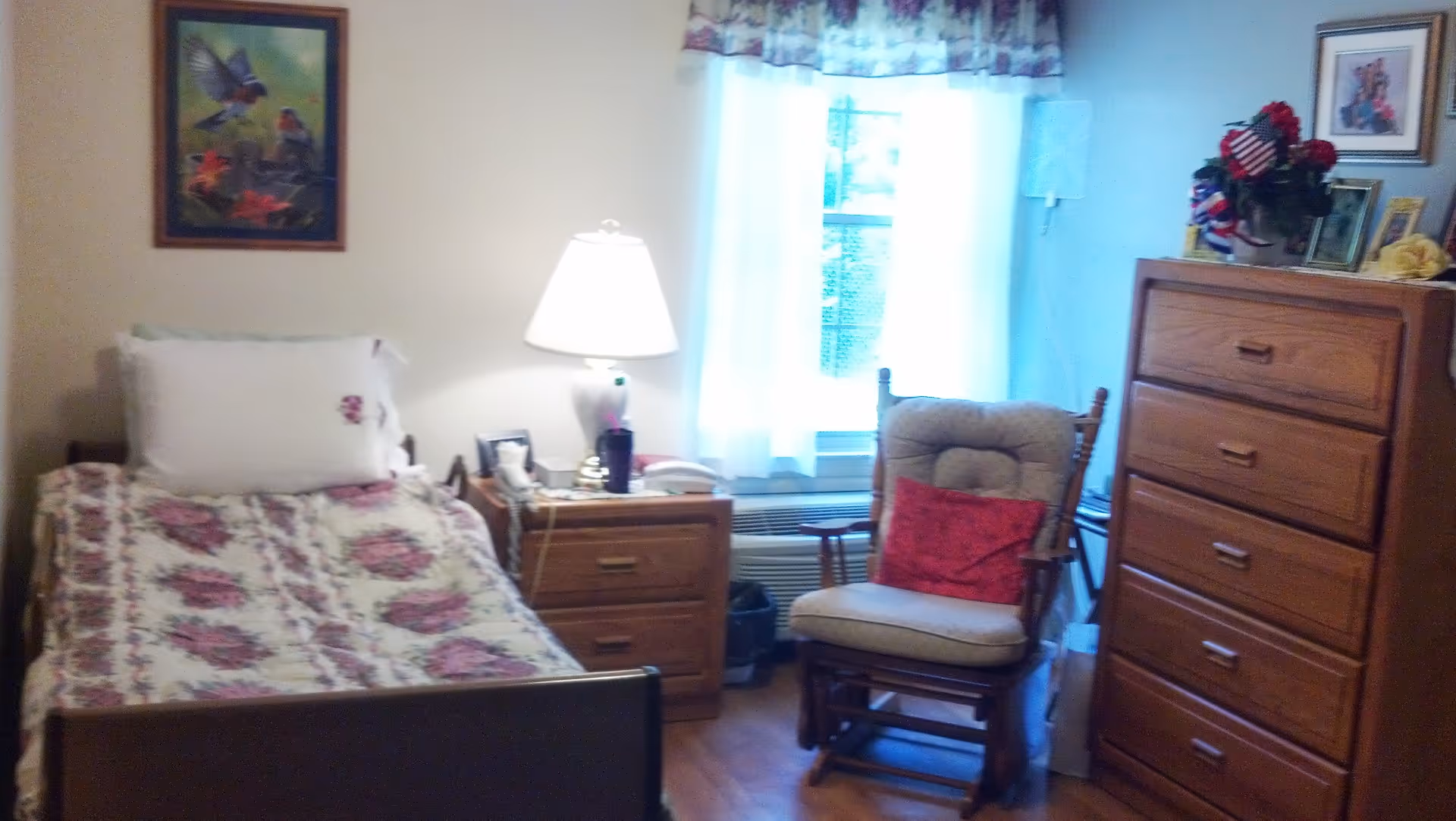 A cozy senior living bedroom with a single bed covered in a floral quilt, a wooden nightstand with a lamp, a telephone, and other small items. Next to the nightstand is a cushioned wooden rocking chair with a red pillow. A window with sheer curtains and a floral valance lets in natural light. A tall wooden dresser with framed photos and a flower arrangement is on the right side of the room. A framed picture of birds hangs on the wall above the bed.