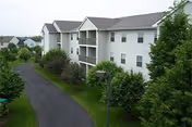 Exterior view of a multi-story residential building with white siding and multiple balconies, surrounded by green trees and a paved driveway leading up to the building.