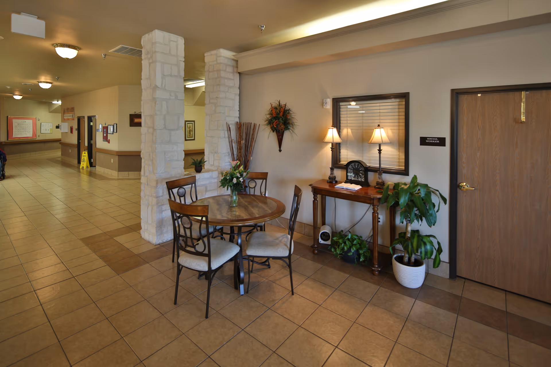 Interior view of a nursing and rehabilitation center hallway with a small round wooden table and four chairs. The table has a vase with flowers. There are two stone pillars next to the table. On the right side, there is a wooden console table with two lamps, a clock, and some plants underneath. A door labeled 'Social Worker's Office' is visible next to the console table. The hallway has tiled floors and beige walls with ceiling lights.