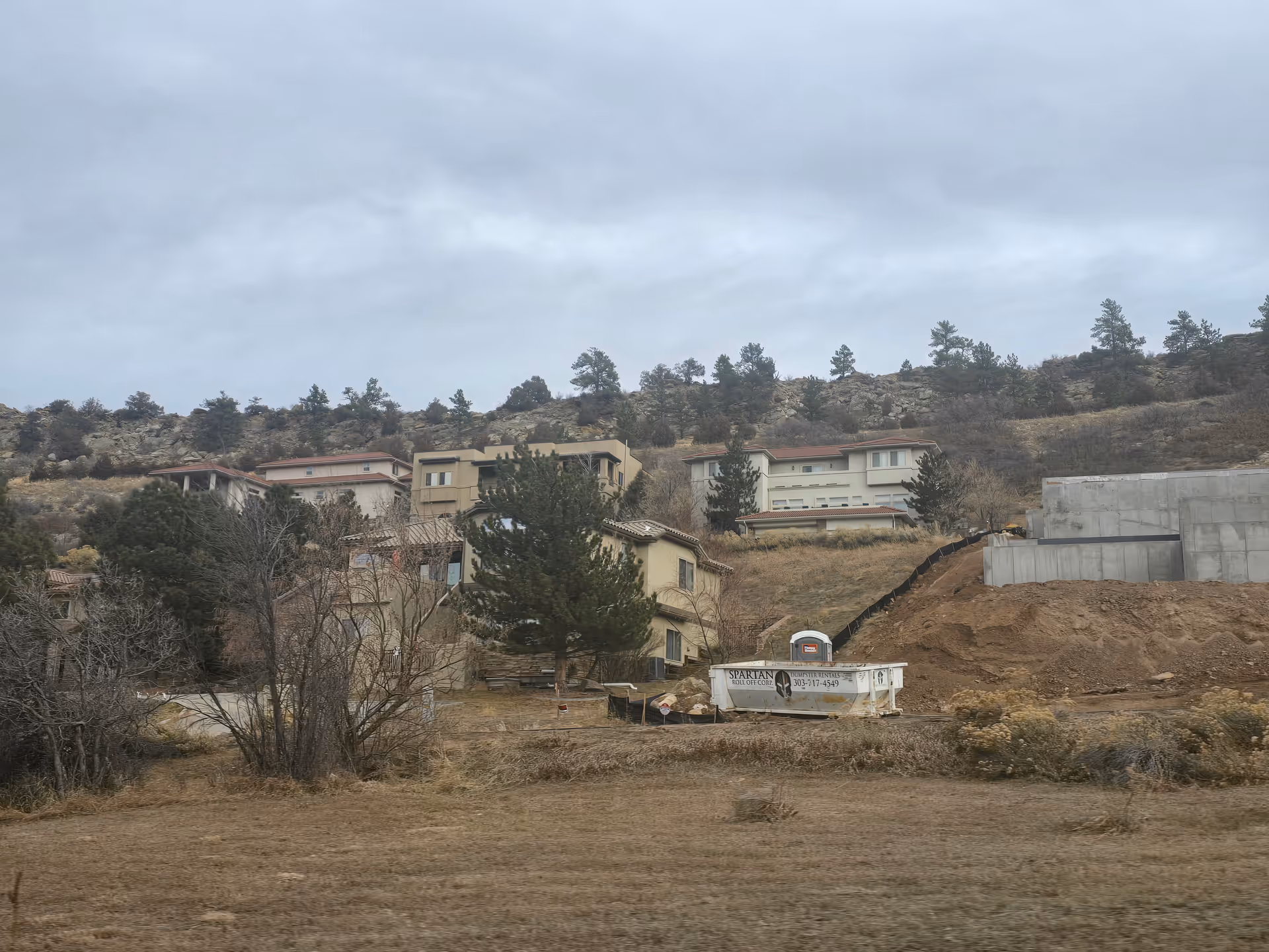 Hillside view with several houses and a construction site featuring a dumpster and concrete foundation. Trees and dry grass cover the area under a cloudy sky.