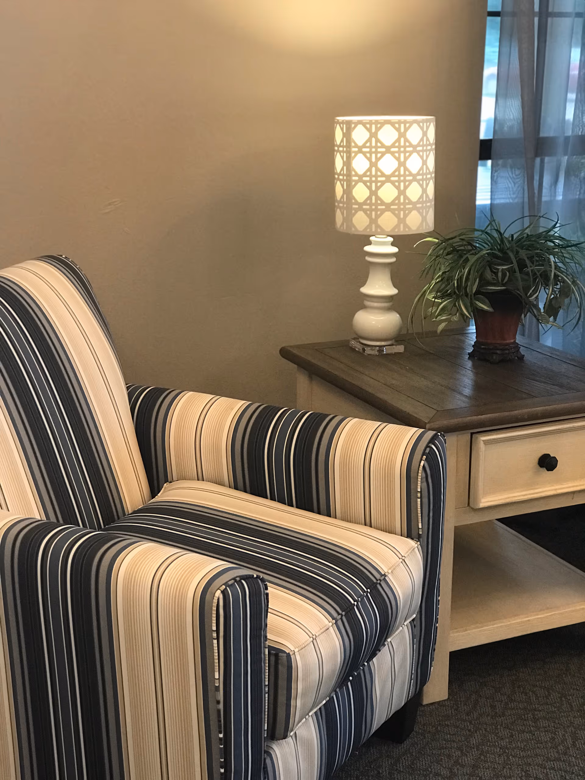 A cozy corner featuring a striped armchair with blue, beige, and white colors next to a wooden side table. On the table, there is a white decorative lamp with a patterned shade and a potted green plant. Sheer curtains cover a window in the background.