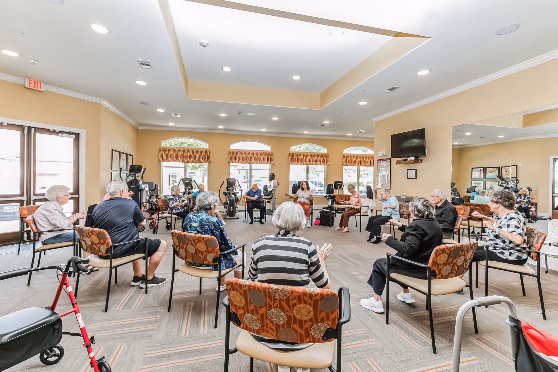 A group of elderly people seated in a circle in a spacious, well-lit room with beige walls and large windows. They appear to be participating in a group activity or exercise session. The room has exercise equipment along the walls and a large mirror on one side. There are walkers and mobility aids visible near some chairs.