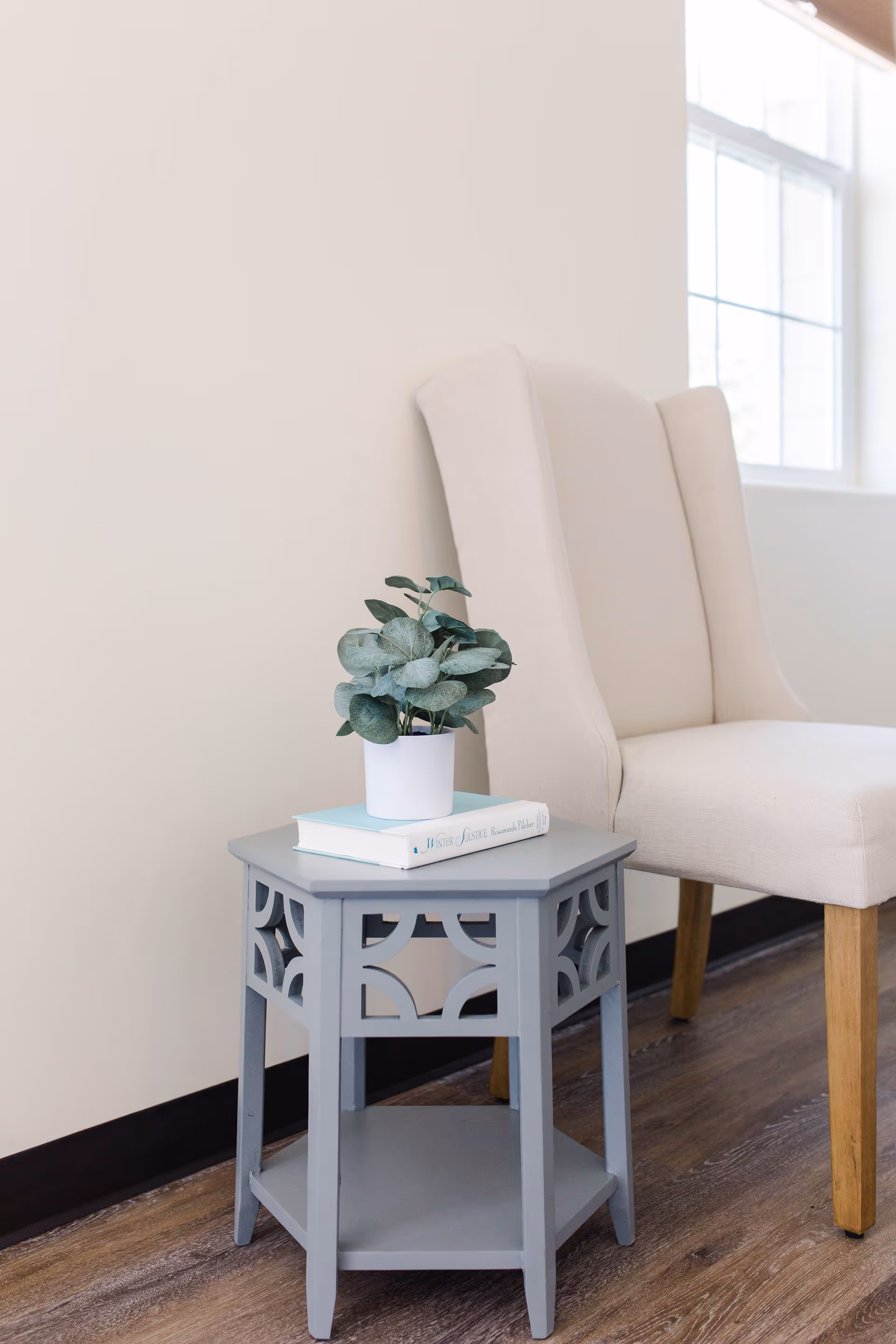 A beige upholstered chair next to a small gray decorative side table with a potted plant and a book on top, set against a light-colored wall and a window in the background.