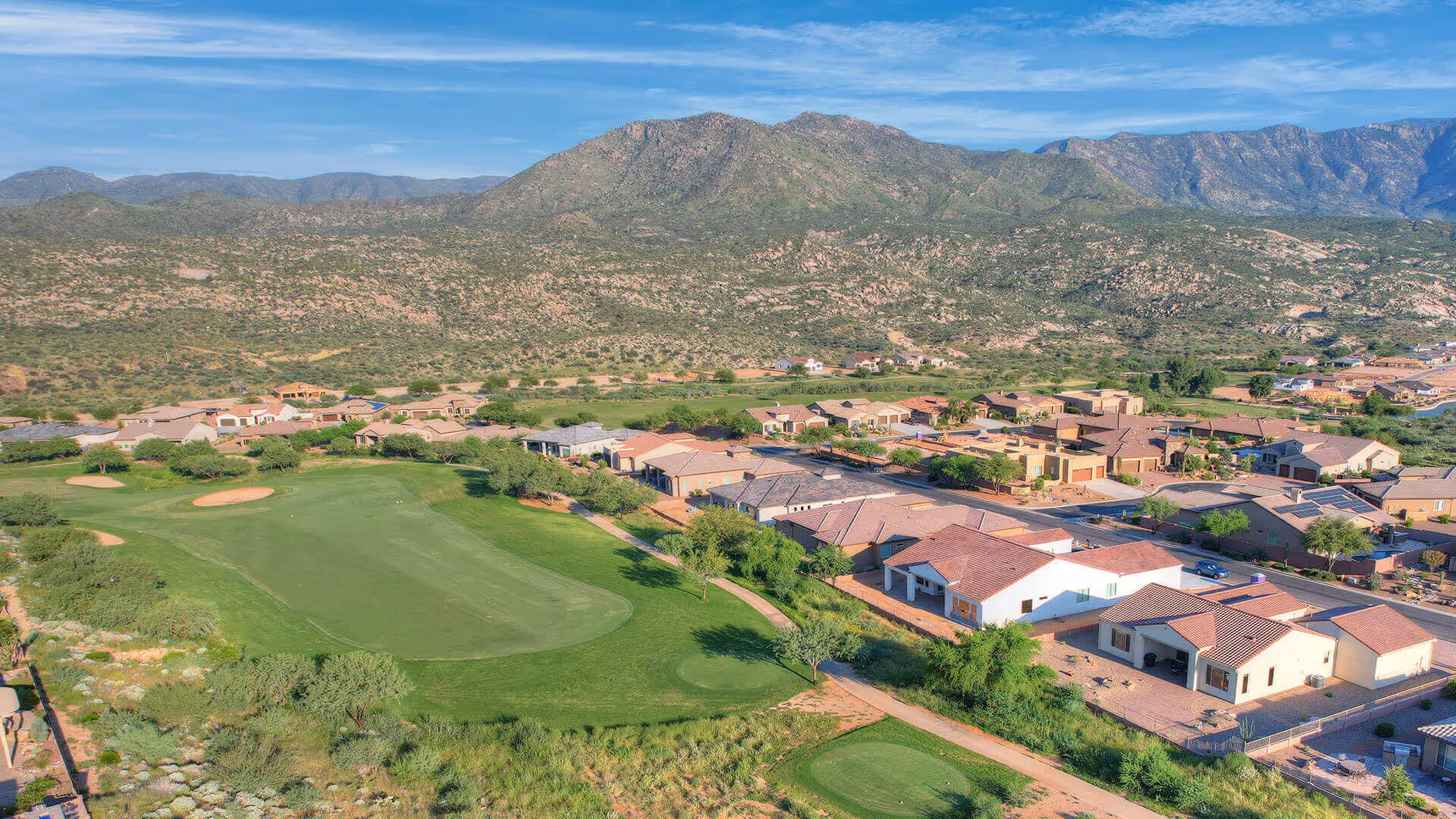 Aerial view of a golf course and adjacent single-story homes with mountains in the background.