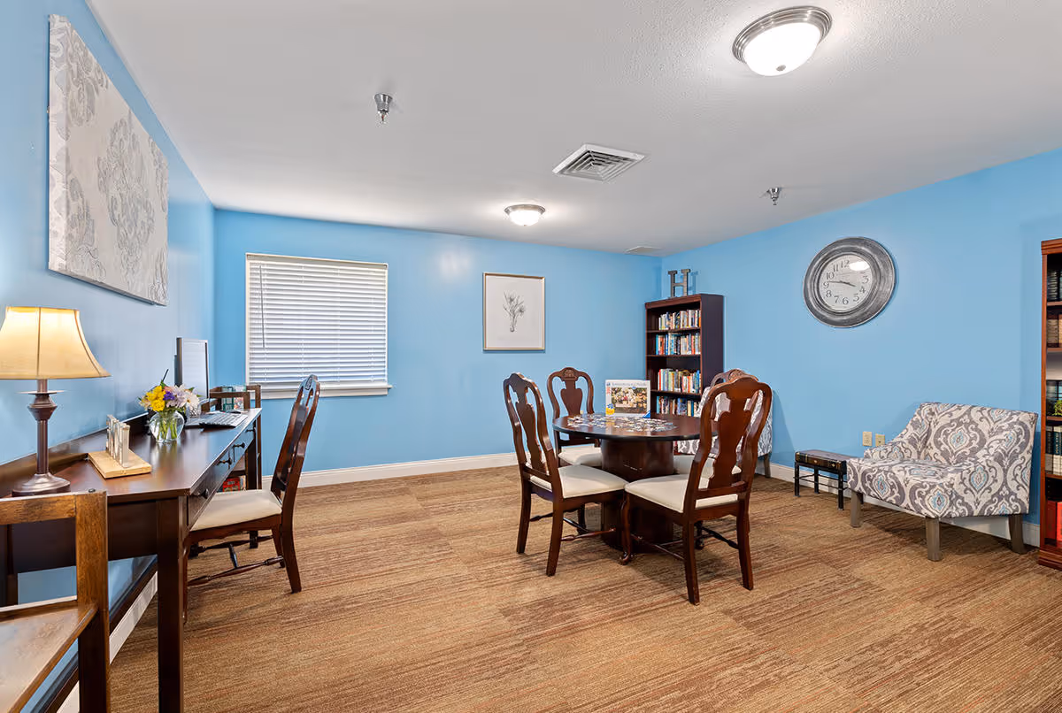 A cozy room with light blue walls and brown carpet featuring a round wooden table with four chairs, a patterned armchair, a bookshelf filled with books, a wall clock, and a desk with a lamp, flowers, and a computer. The room is well-lit with ceiling lights and has framed artwork on the walls.