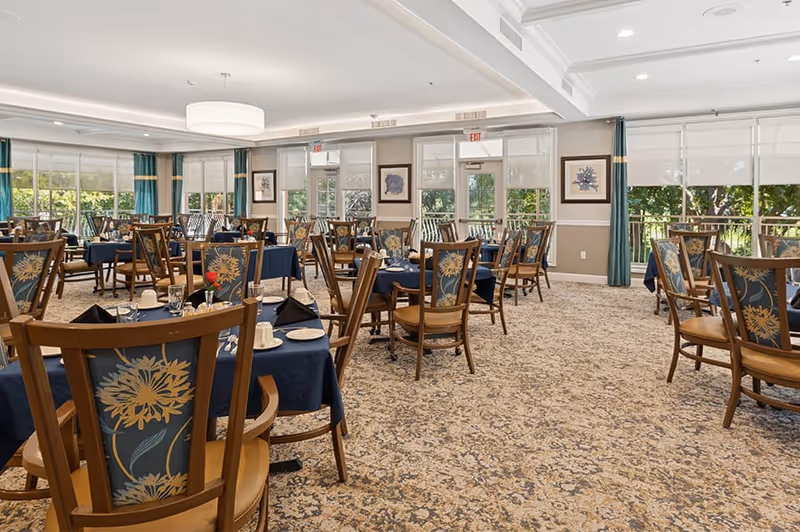 A spacious dining room with multiple wooden tables and chairs arranged neatly. The tables are covered with dark blue tablecloths and set with white plates, cups, and silverware. The chairs have floral patterned upholstery. Large windows with white blinds and green curtains allow natural light to fill the room, and framed artwork hangs on the walls.