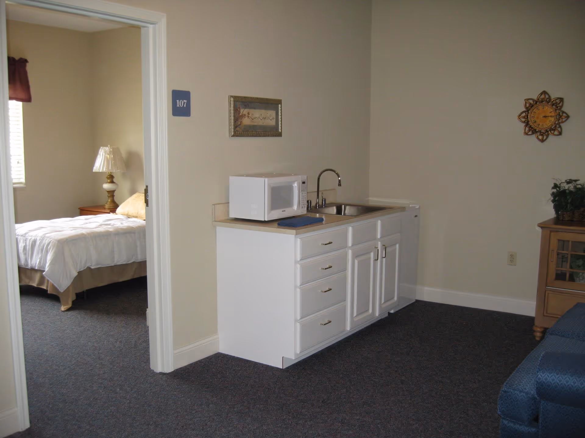 Interior of a small suite showing a kitchenette with sink, microwave and cabinets and an adjacent bedroom with a bed and lamp visible through a doorway.