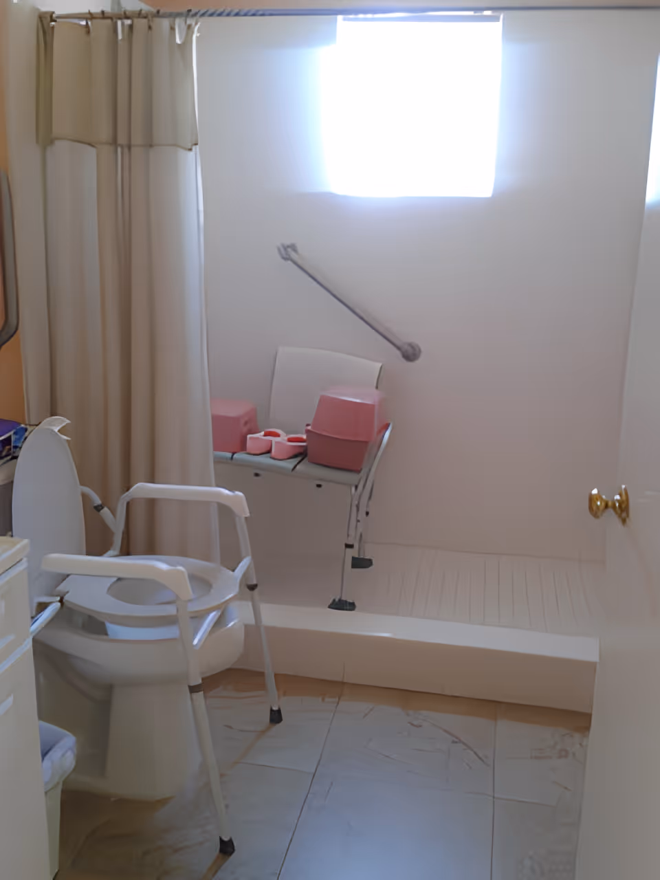 A bathroom equipped for accessibility with a white toilet that has arm supports, a shower area with a white chair and pink bath accessories, a grab bar on the wall, beige shower curtains, and a small window letting in natural light.