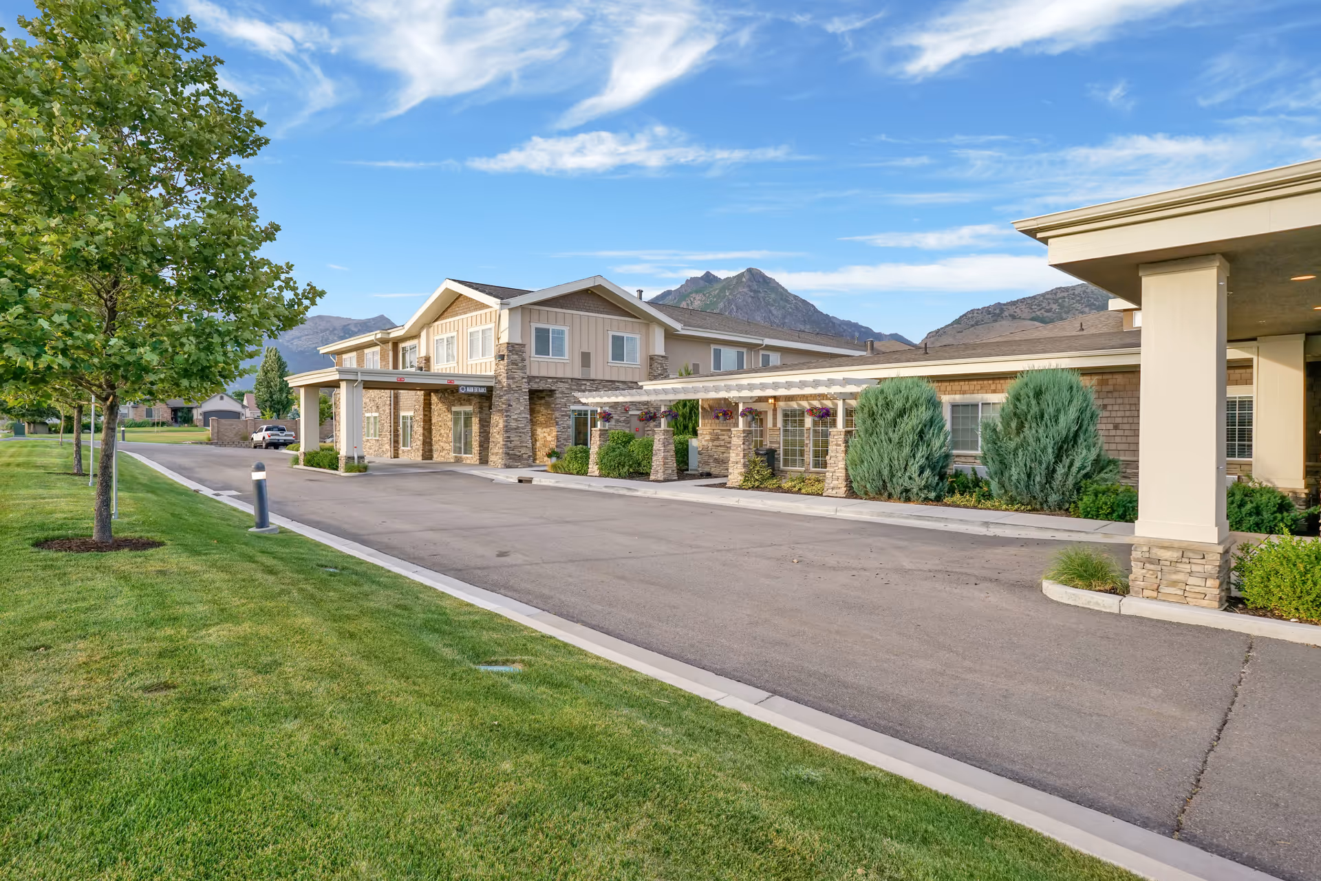 Exterior view of a senior living facility named Highland Glen with a driveway, green lawn, trees, and mountains in the background under a partly cloudy sky.
