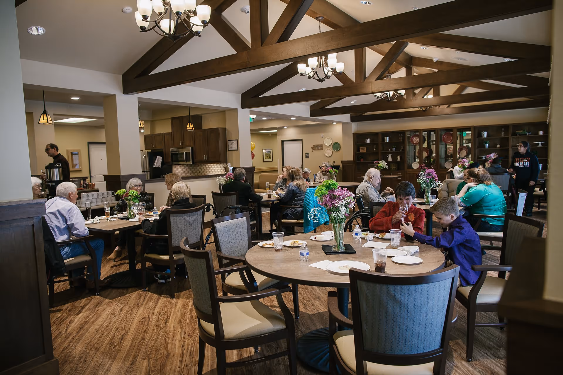 A dining room in a senior living facility with several elderly people and a child seated at round and rectangular tables. The room has wooden floors, exposed wooden beams on the ceiling, chandeliers, and a kitchen area in the background. Tables are decorated with vases of flowers and have plates, cups, and water bottles on them.