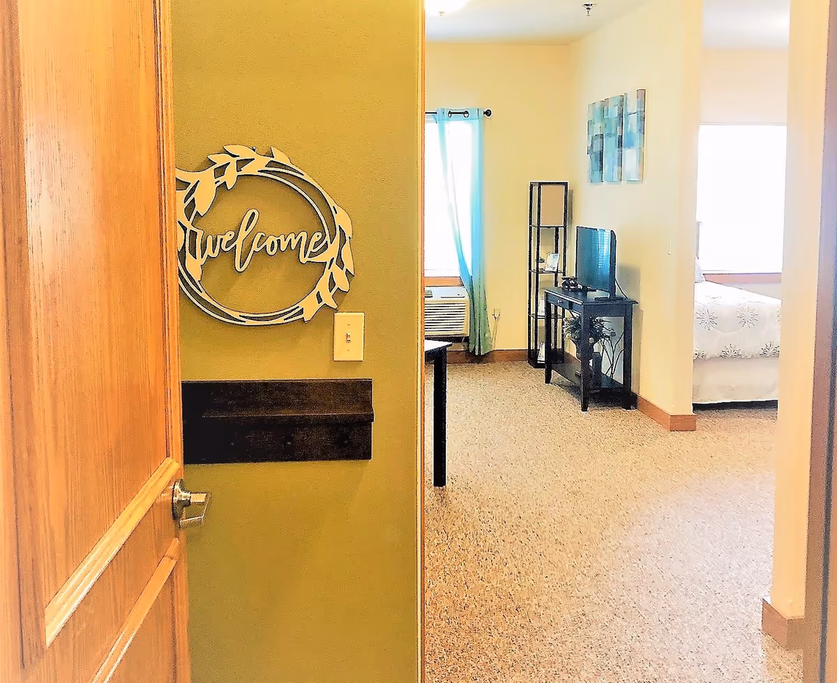 View through an open wooden door into a warmly lit interior space with beige walls and carpet. A decorative 'welcome' sign is mounted on the wall near the door. Inside, there is a small living area with a TV on a stand, a tall shelving unit, and a window with blue curtains. A bedroom with a bed covered in white bedding with a subtle pattern is visible through an open doorway.