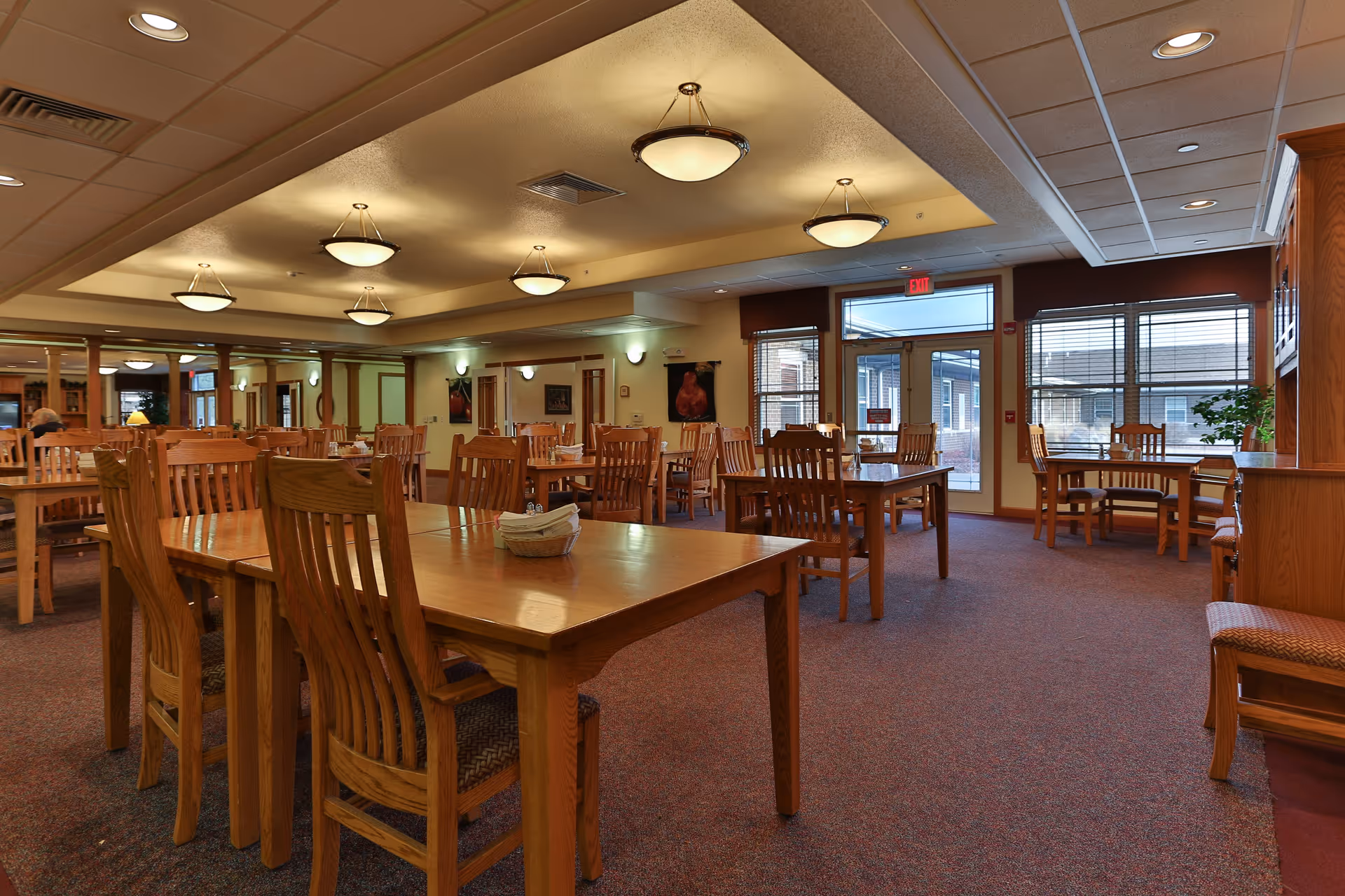 A spacious dining room with multiple wooden tables and chairs arranged neatly. The room has a carpeted floor, ceiling lights, large windows with blinds, and a glass door leading outside. The walls are decorated with framed pictures and there is a wooden cabinet on the right side.