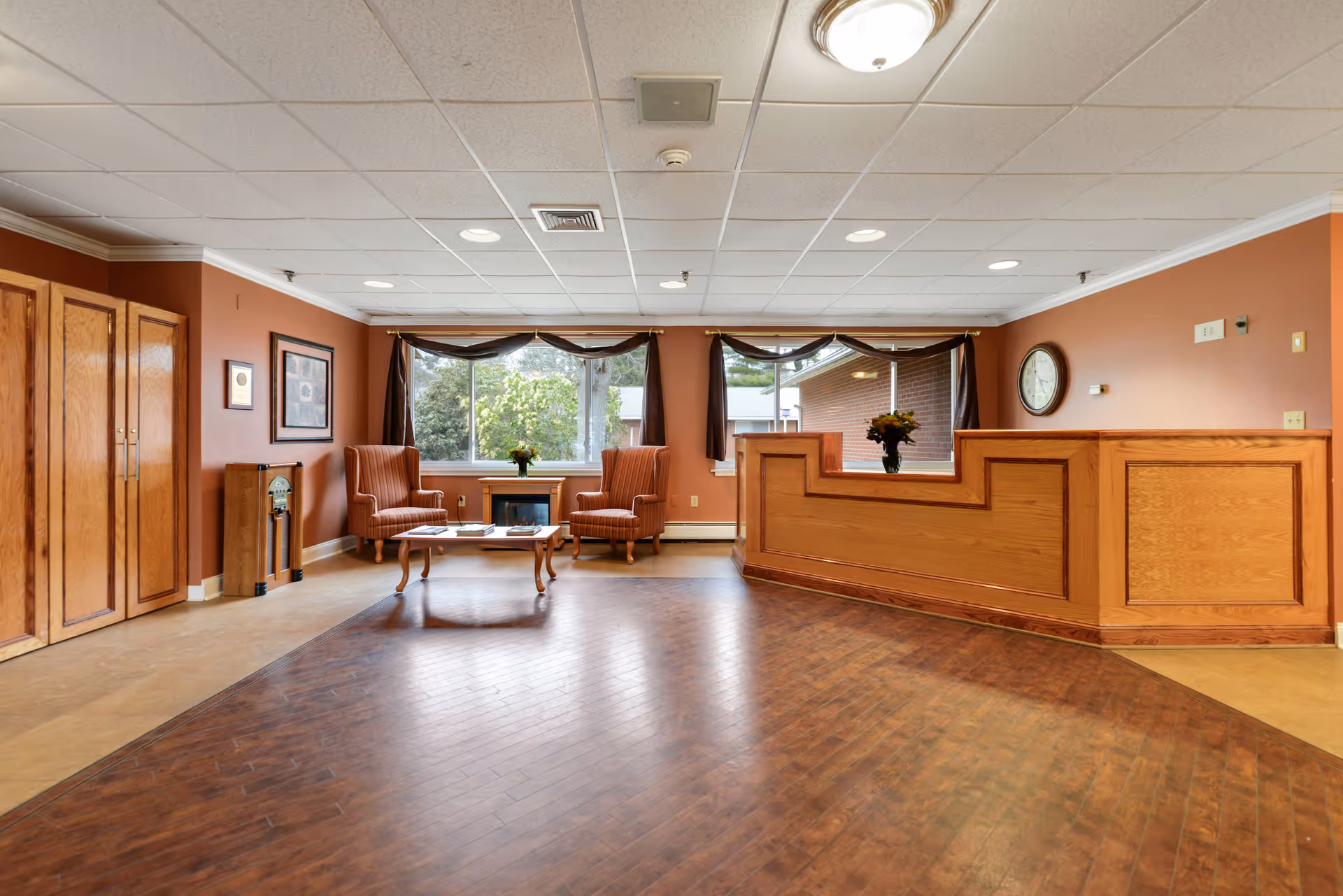 Reception area with a wooden front desk on the right, two orange armchairs and a coffee table with flowers in front of large windows with dark curtains, wooden cabinets on the left, and a clock on the wall.