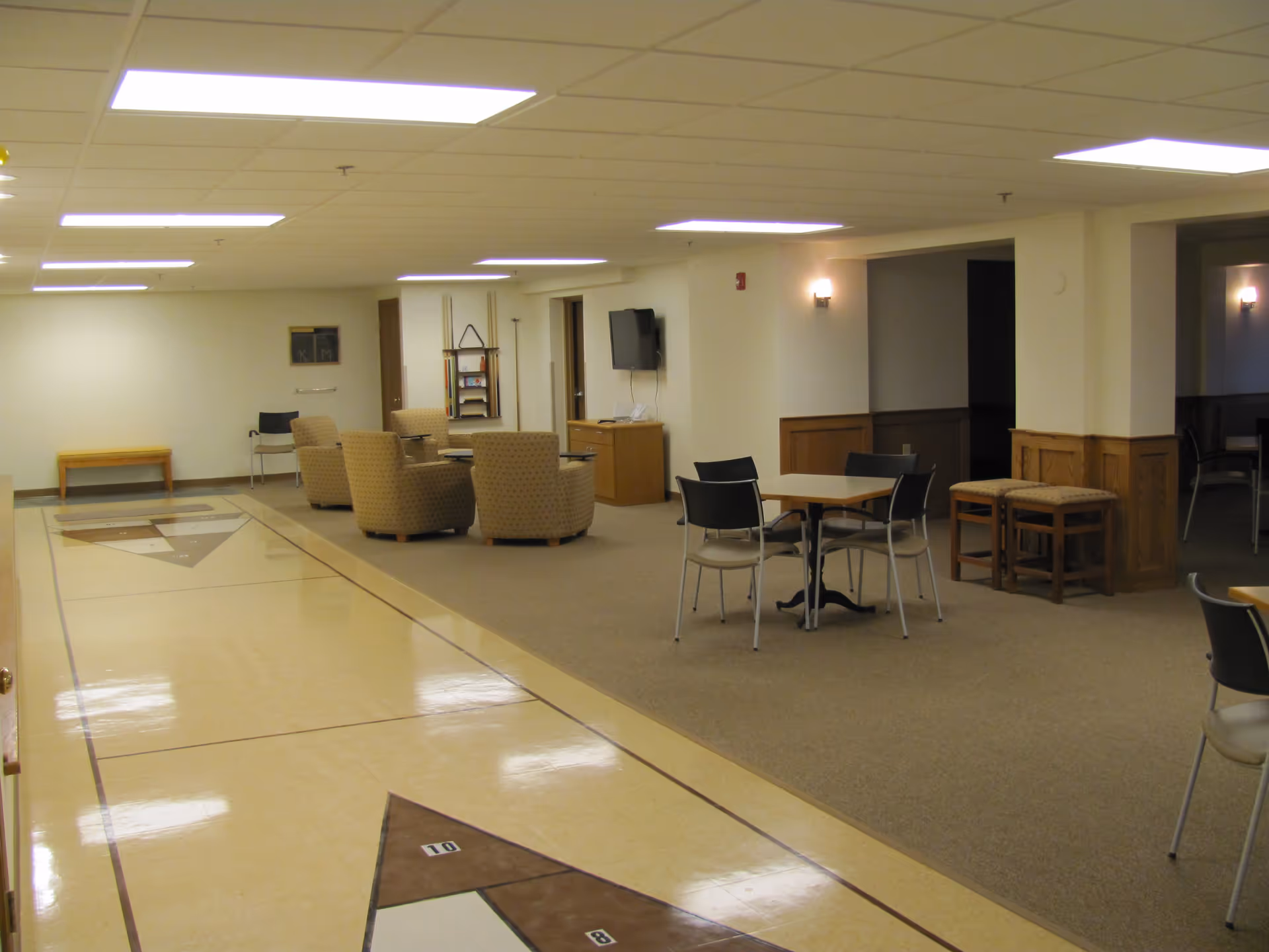 Interior view of a senior living facility common area with a shuffleboard court on the left, several upholstered armchairs arranged around a small table, a wall-mounted TV, and a few tables with chairs on the right side. The room has a drop ceiling with fluorescent lights and beige walls with wooden paneling.