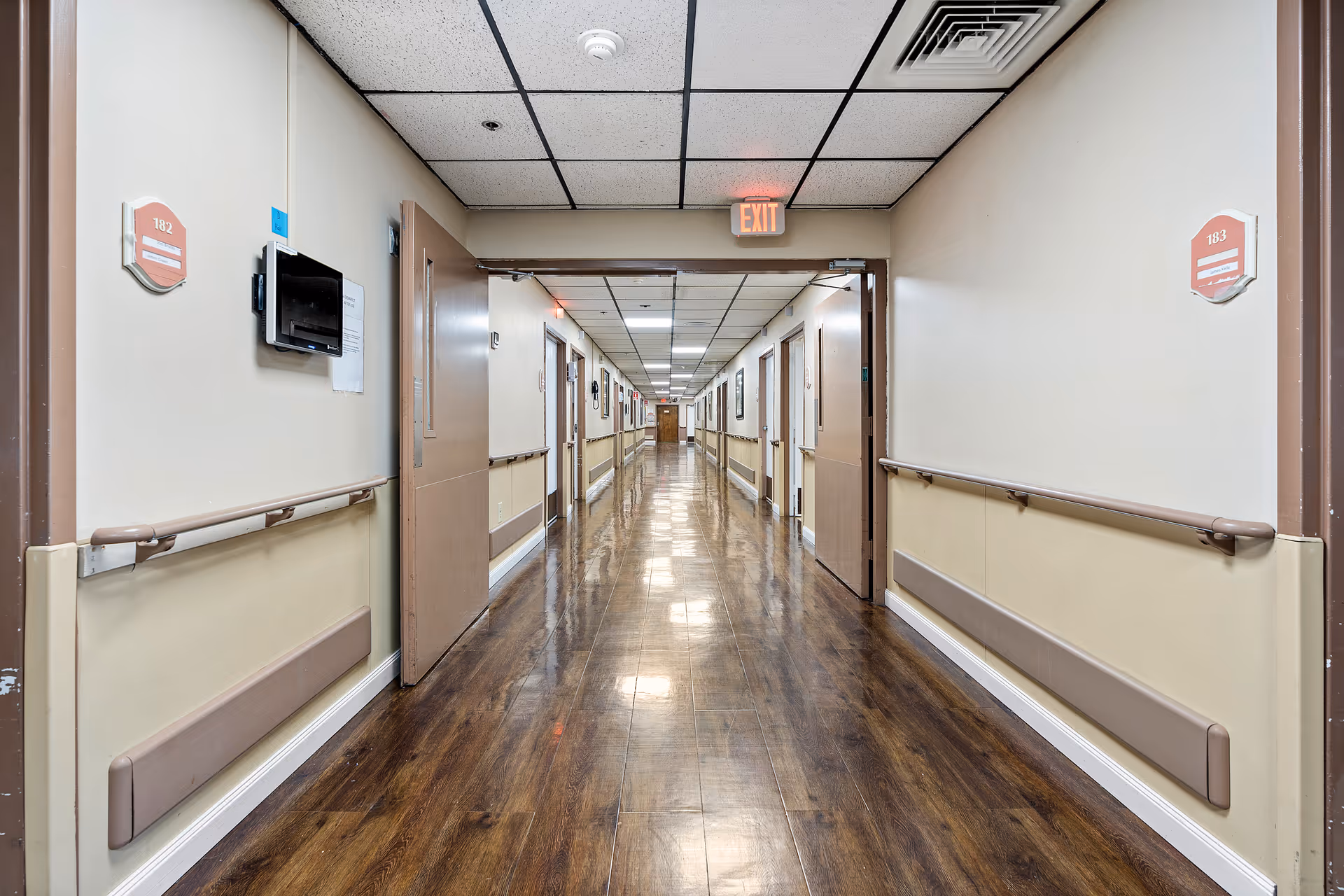 A long, clean hallway in a nursing center with polished wooden floors, beige walls with handrails, and several open doors along the corridor. Room numbers 182 and 183 are visible on the walls near the doors. An exit sign is illuminated at the end of the hallway.