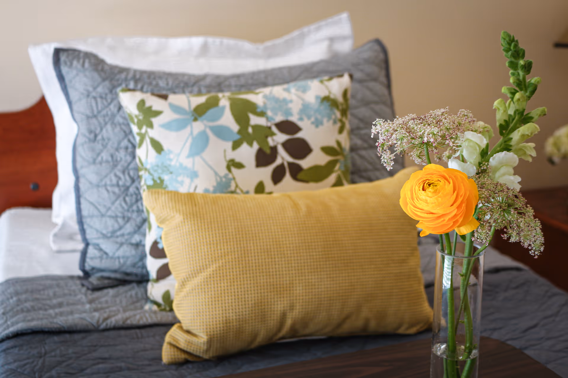 Close-up of a bed with three decorative pillows in different patterns and colors, including a yellow pillow in front, a floral pillow in the middle, and a gray quilted pillow at the back. A clear glass vase with a yellow flower and other small flowers is placed on a wooden surface next to the bed.
