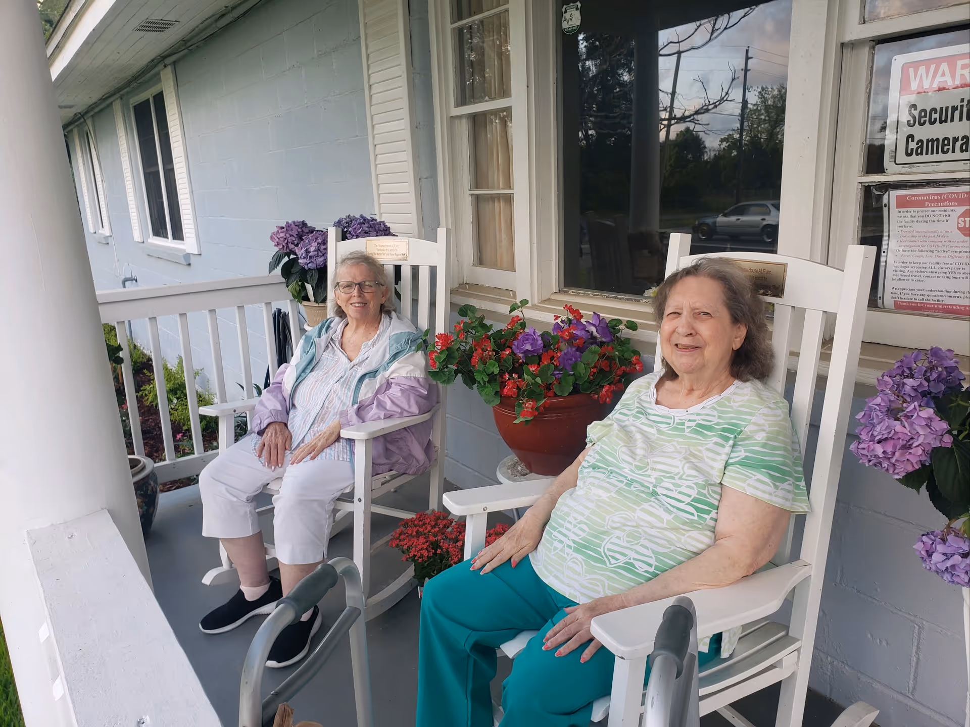 Two elderly women sitting on white rocking chairs on a porch of a building with light blue walls. There are potted flowers with purple and red blooms placed between and beside them. A window and a door with security camera and COVID-19 precaution signs are visible behind them.
