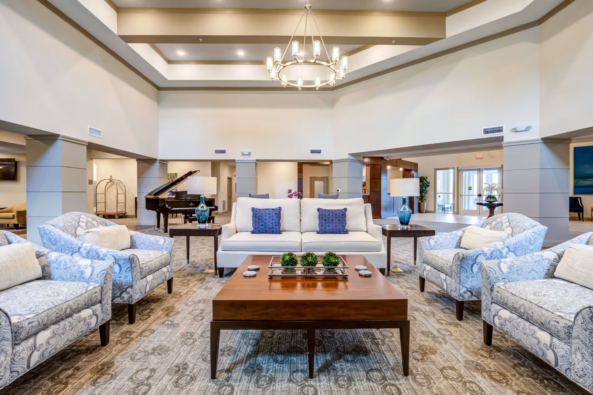 Spacious and well-lit senior living facility common area with a white sofa adorned with two blue patterned pillows, four matching armchairs with light blue and white paisley patterns, a wooden coffee table with decorative greenery, two side tables with blue glass lamps, a grand piano in the background, and a chandelier hanging from the high ceiling.