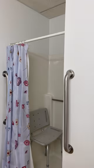 A shower area with a floral-patterned shower curtain partially open, revealing a white shower chair inside. There are two stainless steel grab bars mounted on the walls for support and safety.