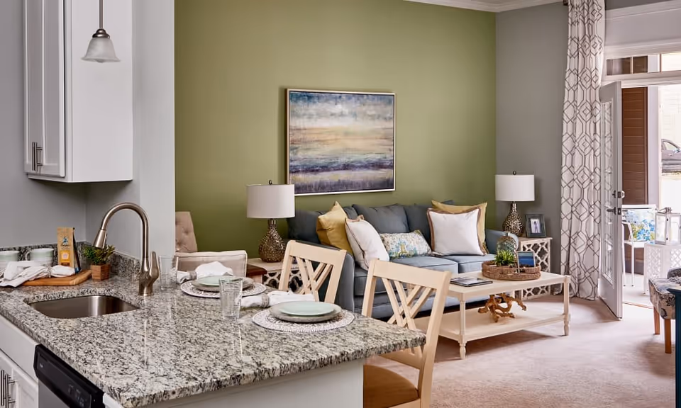 Open-concept living area with a granite kitchen island and place settings in the foreground and a sofa against a green accent wall with a coffee table and lamps in the background.