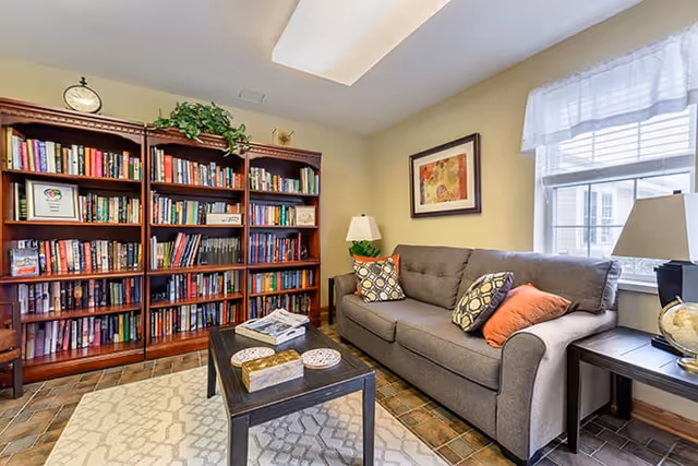 A cozy living room with a gray sofa adorned with patterned and solid orange pillows, a wooden coffee table with decorative items and magazines, a large wooden bookshelf filled with books, a framed artwork on the wall, two table lamps, and a window with white curtains letting in natural light.
