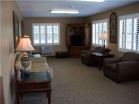 Cozy senior living common room with sofa, chairs, lamps, windows, and a TV cabinet.