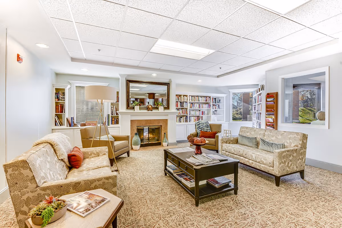 A bright and cozy living room with two patterned sofas, two armchairs, a dark wooden coffee table with magazines and decorative items, a fireplace with a large mirror above it, built-in bookshelves filled with books, and large windows letting in natural light.