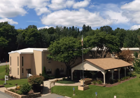 Exterior view of a beige two-story building with a covered entrance and a flagpole in front, surrounded by green trees and a well-maintained lawn under a partly cloudy sky.