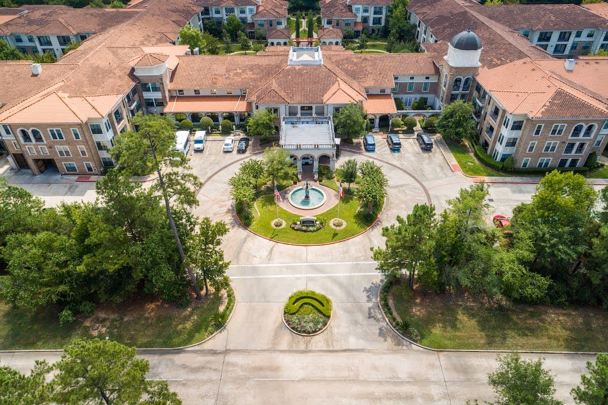 Aerial view of Conservatory At Alden Bridge senior living facility showing a large building with terracotta roof tiles, a circular driveway with a central fountain, landscaped greenery, and parked cars.