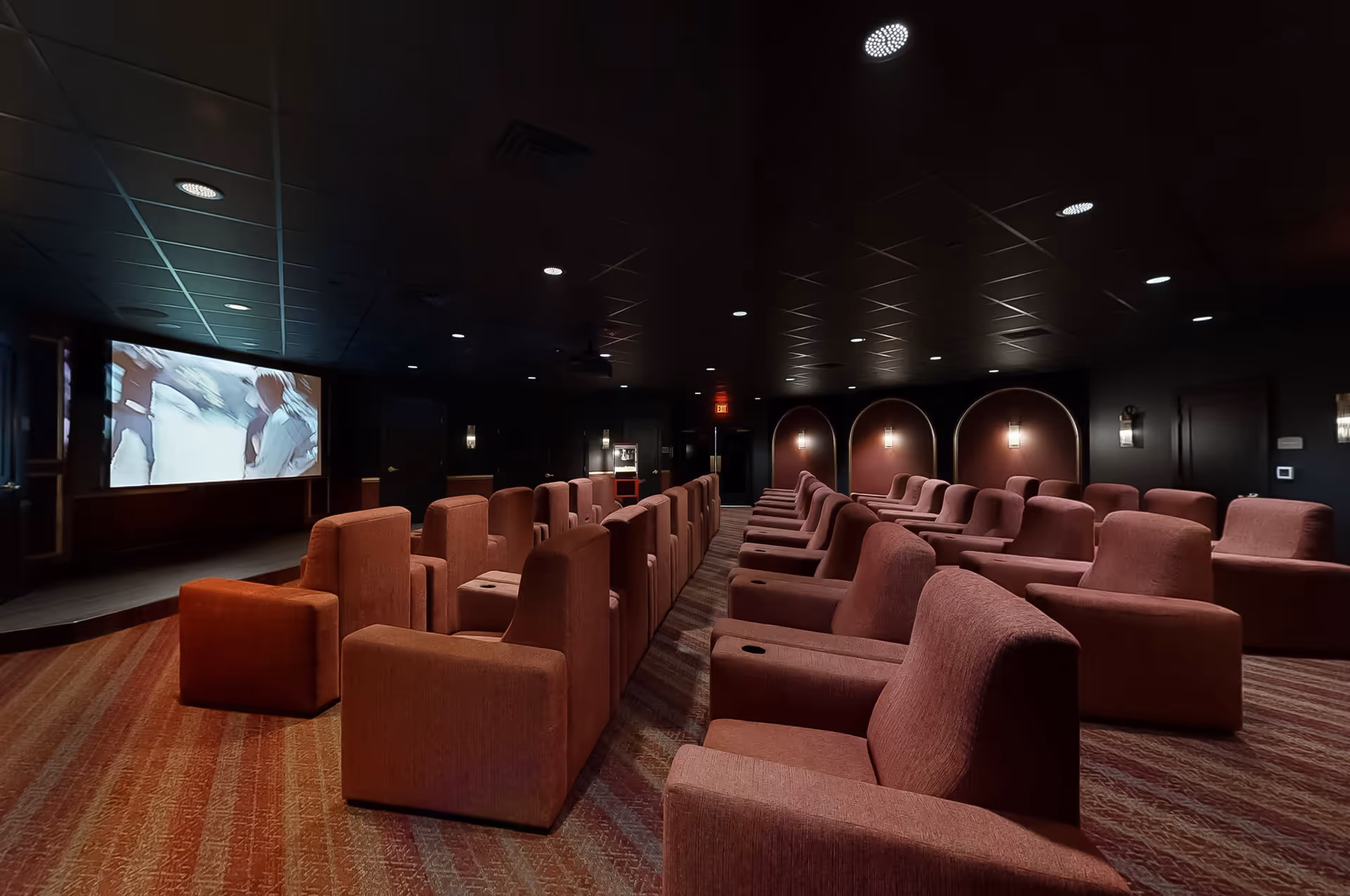Interior of a small movie theater with rows of comfortable, upholstered armchairs facing a large screen showing a movie. The room has dim lighting with wall sconces and a patterned carpet.