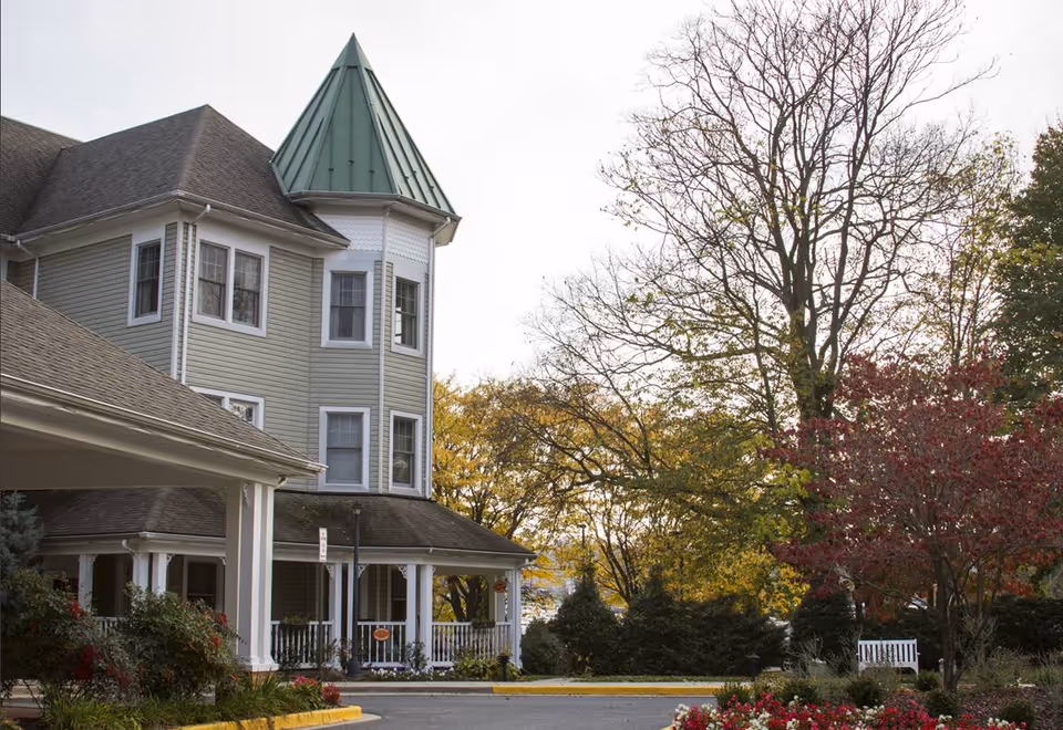 Exterior view of a senior living facility building with a green pointed roof tower, surrounded by trees with autumn foliage and landscaped bushes and flowers. There is a covered entrance and a white bench near the garden area.