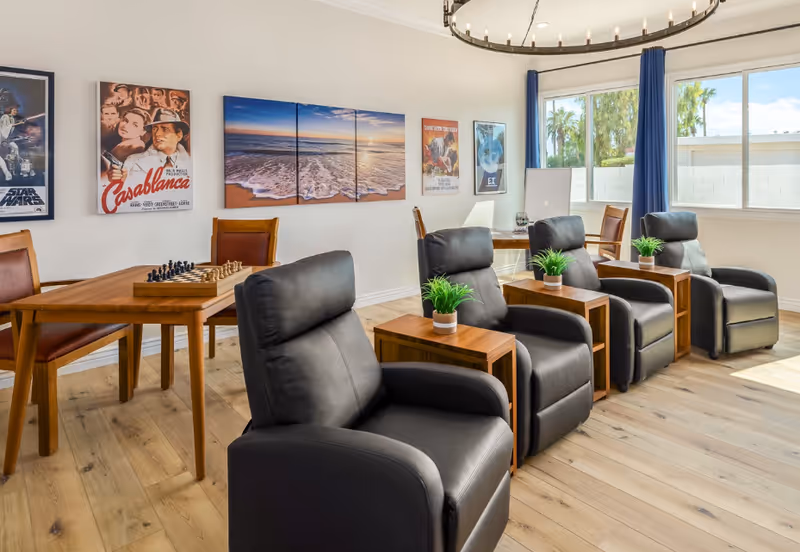 A bright and cozy common area with four black leather recliners each paired with a small wooden side table holding a small green plant. Behind the recliners is a wooden table with a chessboard and four brown leather chairs. The walls are decorated with framed posters including Casablanca, Star Wars, and other classic movies. Large windows with blue curtains let in natural light and show palm trees outside.