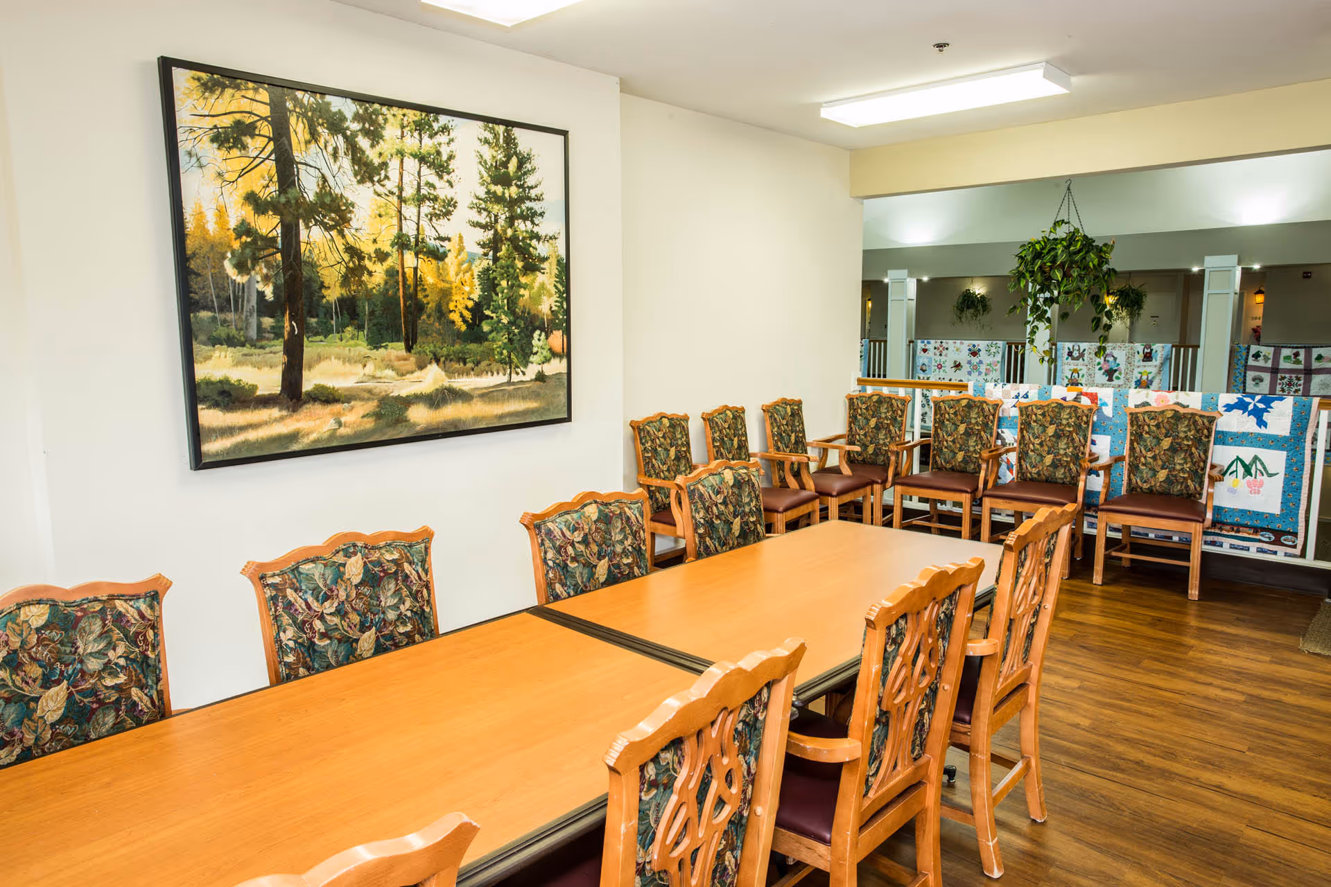 A well-lit room with a long wooden table surrounded by wooden chairs with floral upholstery. On the wall above the table is a large framed painting of a forest scene with tall trees and sunlight filtering through. In the background, there are additional chairs lined up against a railing with colorful quilts hanging behind them, and a hanging plant is visible.
