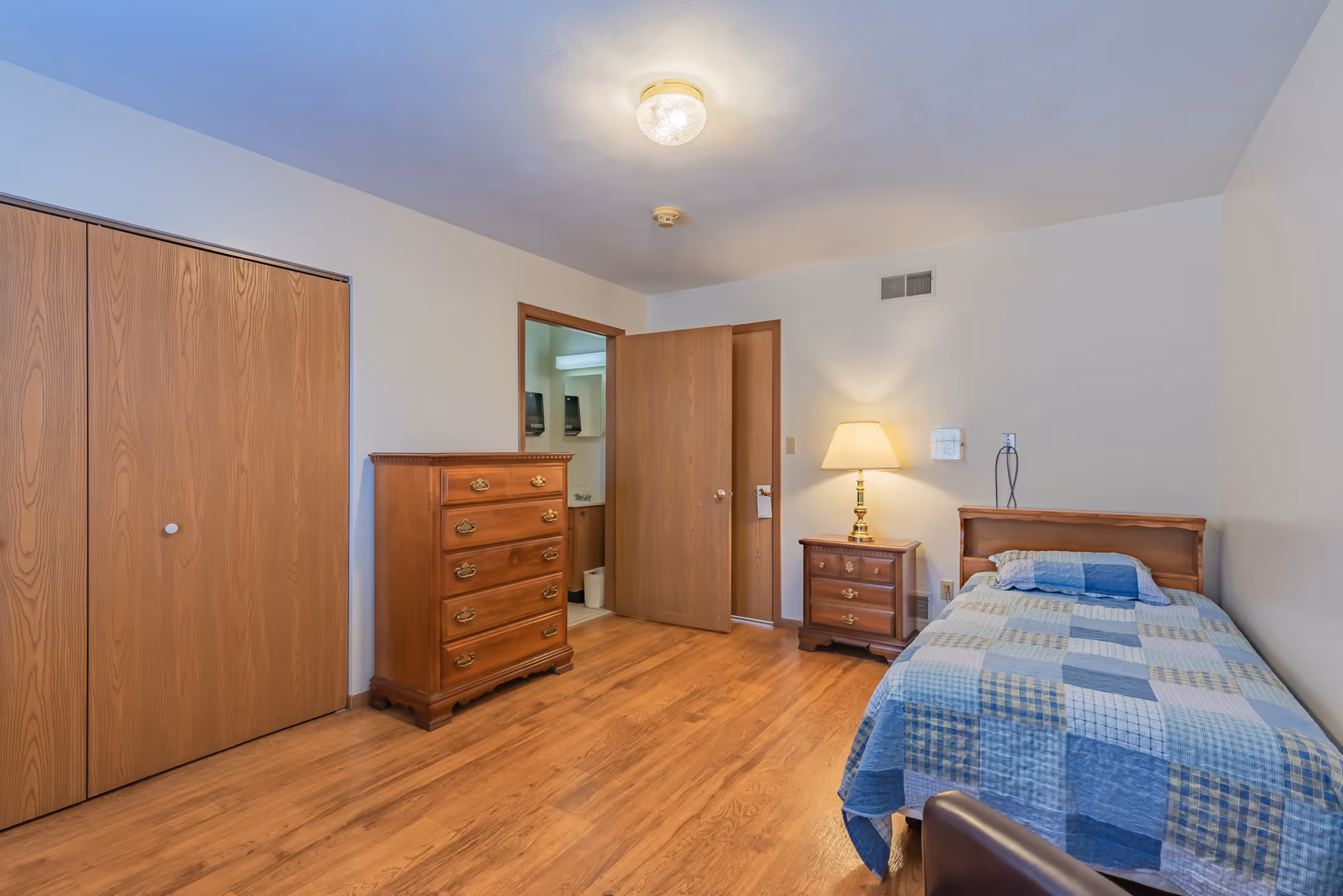 A simple bedroom with wooden flooring, a single bed with a blue and beige patchwork quilt, a wooden nightstand with a lamp, a wooden dresser, and a closet with wooden sliding doors. The door to the room is open, revealing a glimpse of a bathroom.