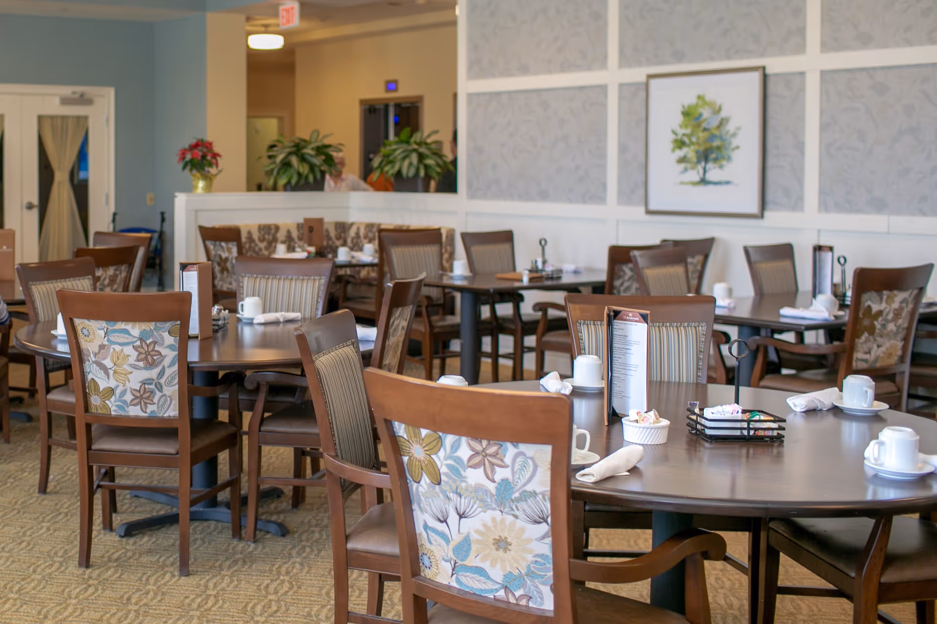 A dining room in a senior living facility with multiple round and rectangular tables set with white cups, napkins, and menus. The chairs have wooden frames with floral and striped upholstery. The room features light-colored walls with framed artwork and potted plants in the background.