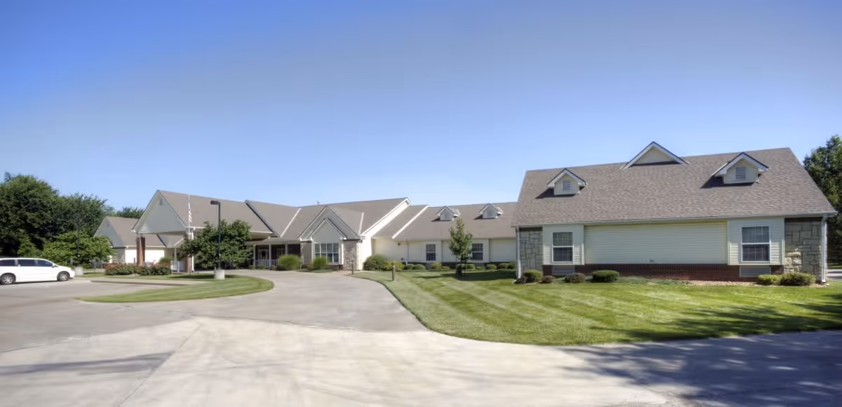 Exterior view of a senior living facility building with a driveway, well-maintained lawn, and clear blue sky. The building has a light-colored facade with multiple roof peaks and dormer windows.