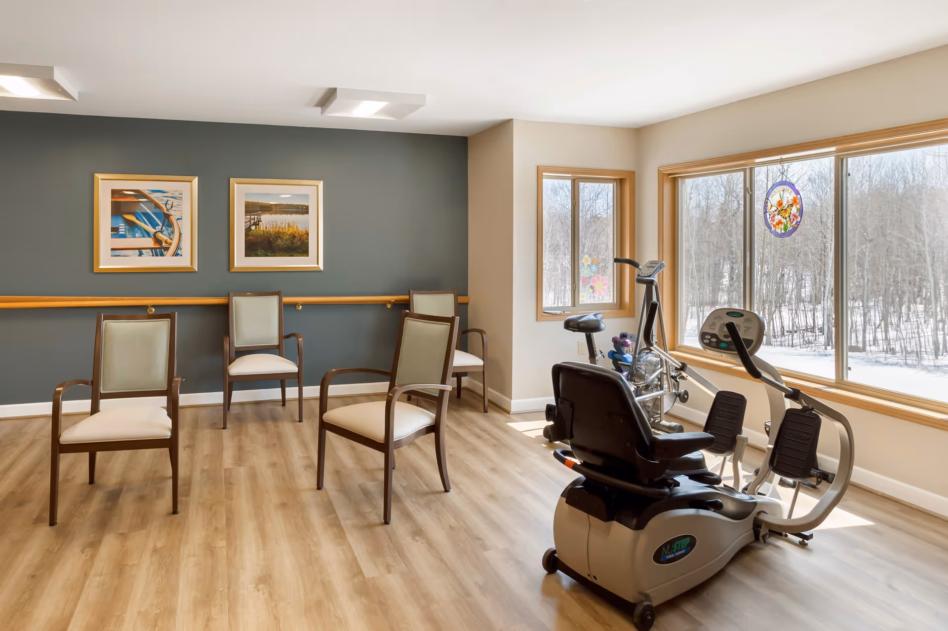 A bright exercise room with large windows showing a snowy outdoor scene. The room has light wood flooring, several wooden chairs with light cushions arranged along the walls, and two exercise bikes positioned near the windows. The walls are painted in neutral tones with two framed pictures hanging on a dark accent wall.