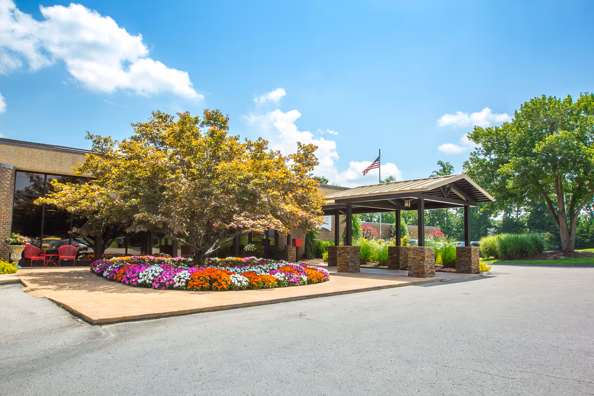 Exterior view of Signature HealthCARE of Putnam County showing the entrance area with a covered driveway, a large tree surrounded by colorful flower beds, and an American flag flying on a flagpole under a bright blue sky with some clouds.