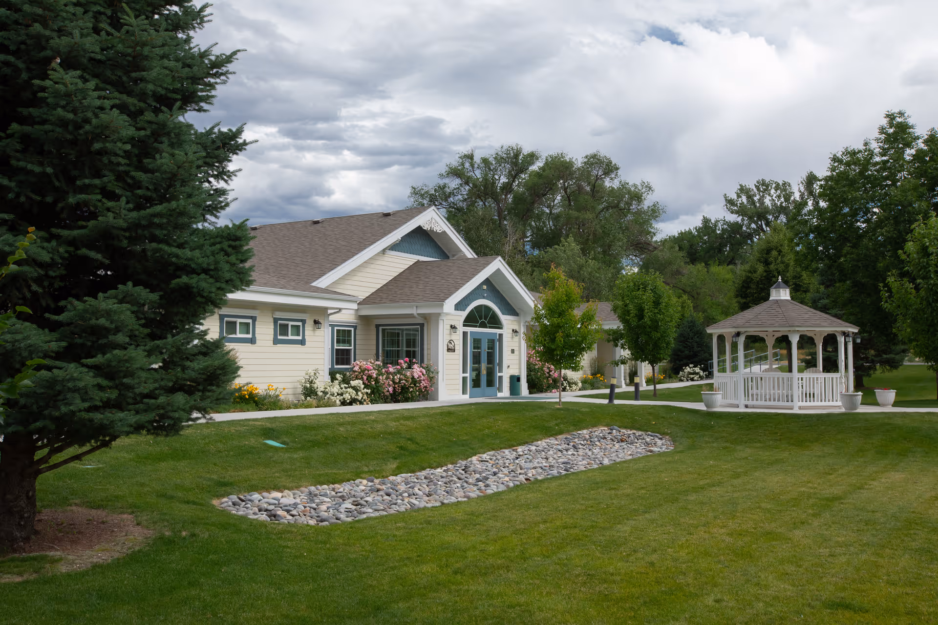 Front of a single-story building with a manicured lawn, gazebo, and trees under a cloudy sky.