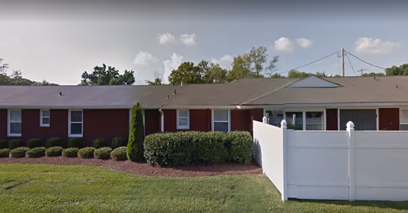 Single-story residential building with red exterior walls, multiple windows, a gray roof, and a white privacy fence on the right side. There are neatly trimmed bushes and green grass in front of the building under a partly cloudy sky.