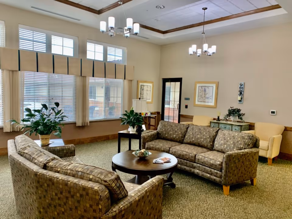 Comfortable senior living lounge with sofas, armchairs, a round coffee table, plants, and large windows.