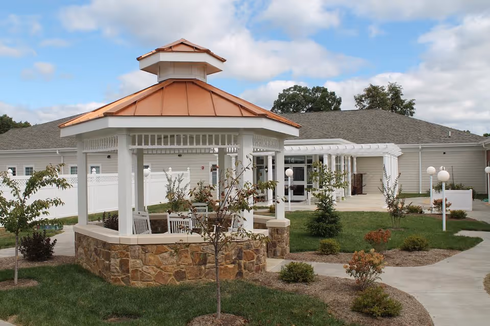 Outdoor area of a nursing home featuring a white gazebo with a copper-colored roof, surrounded by a stone base and white chairs inside. The area has green grass, small trees, shrubs, and paved walkways leading to a building with light-colored siding and a covered entrance. The sky is partly cloudy.